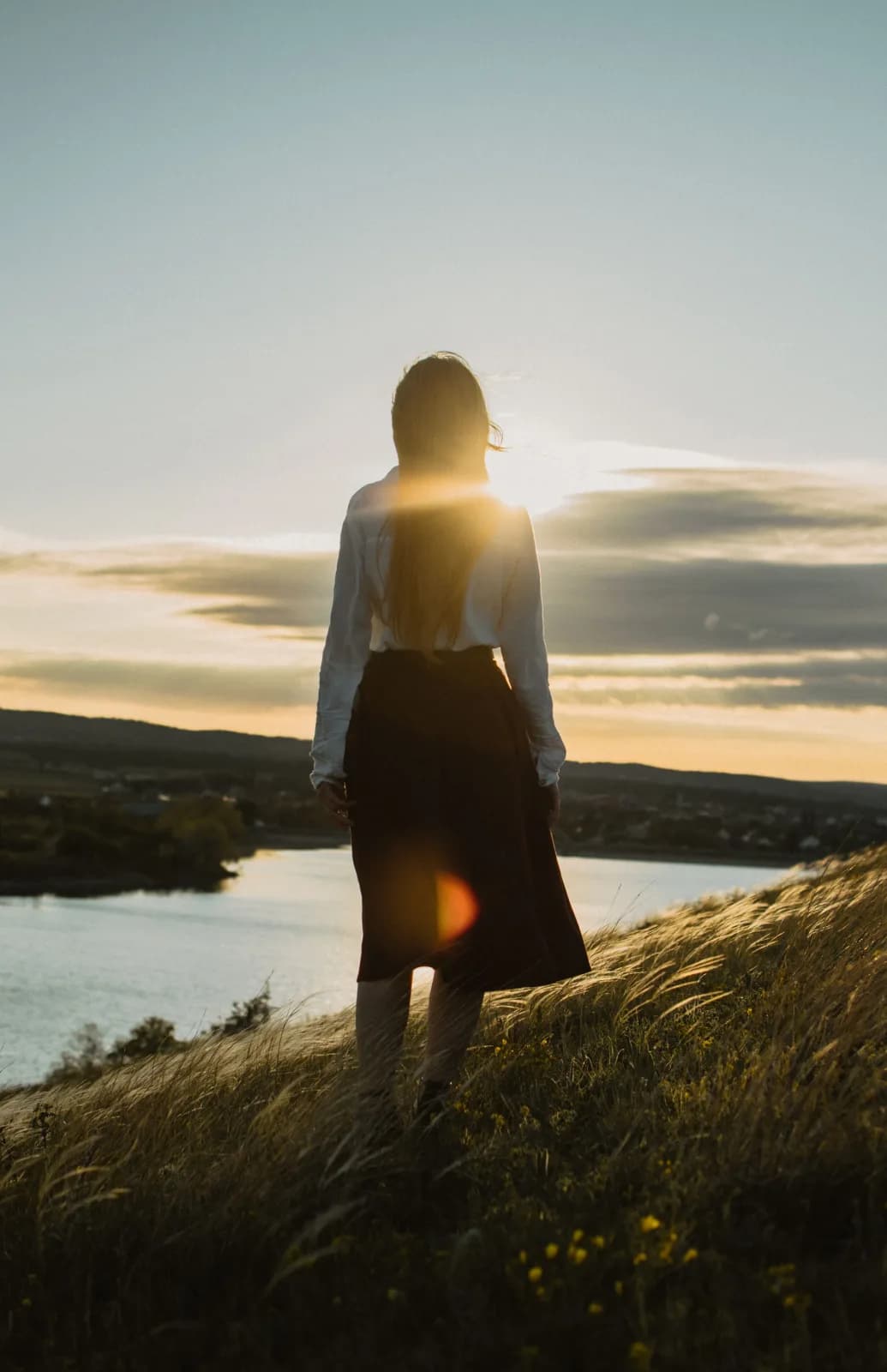 Mujer de espaldas frente a un lago al atardecer, con el sol de frente